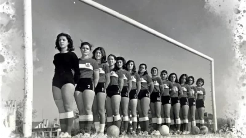 Siyah beyaz tarihi fotoğrafta kalenin önünde yan yana sıralanmış kadın futbol takımı oyuncuları ve futbol topları.