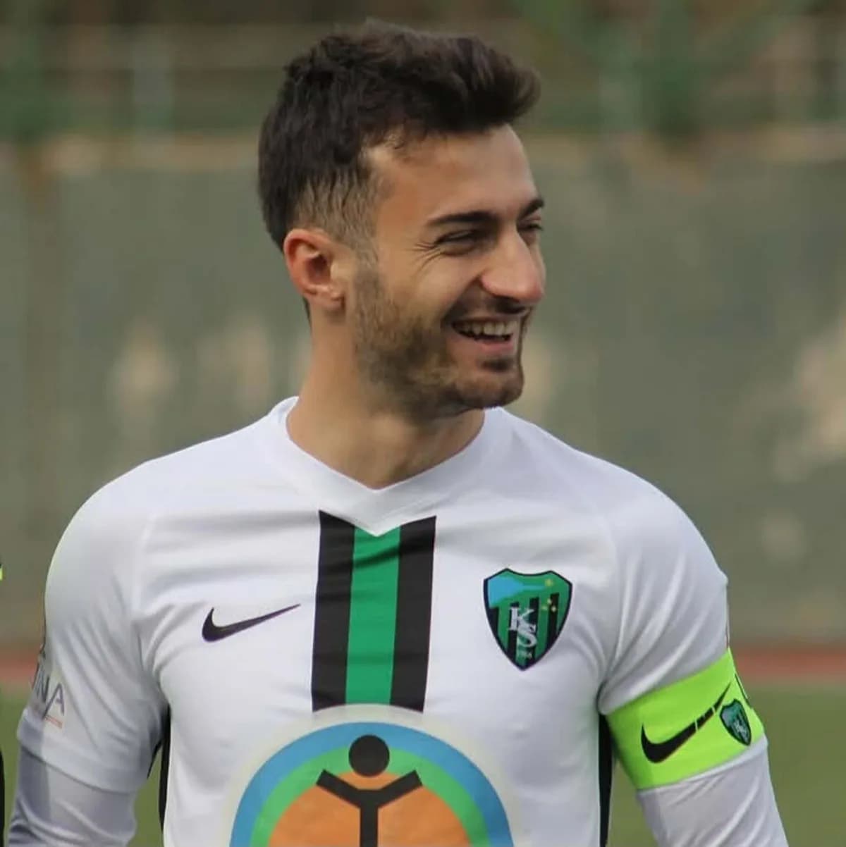 Yeşil-siyah şeritli beyaz forma giyen gülümseyen futbolcu, kolunda sarı kaptan pazubandı