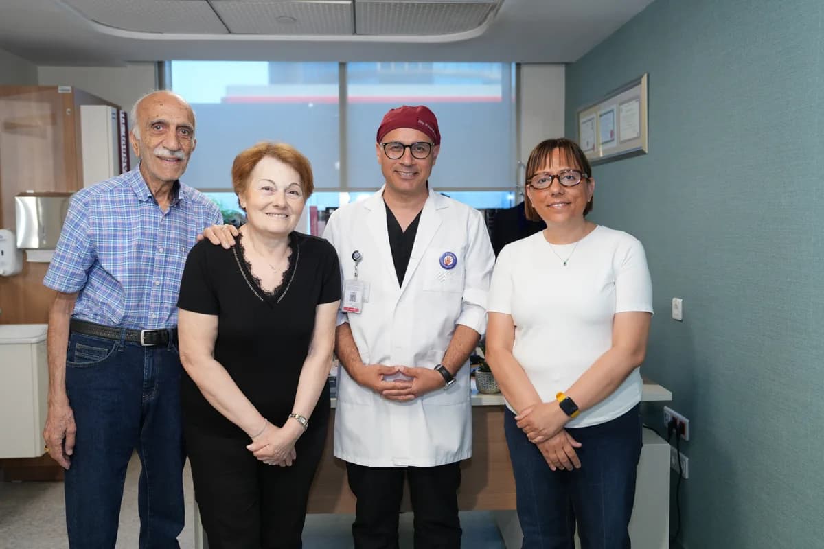 Beyaz önlüklü ve bordo cerrahi bonesi takan doktor, hasta ve iki refakatçiyle muayene odasında poz veriyor