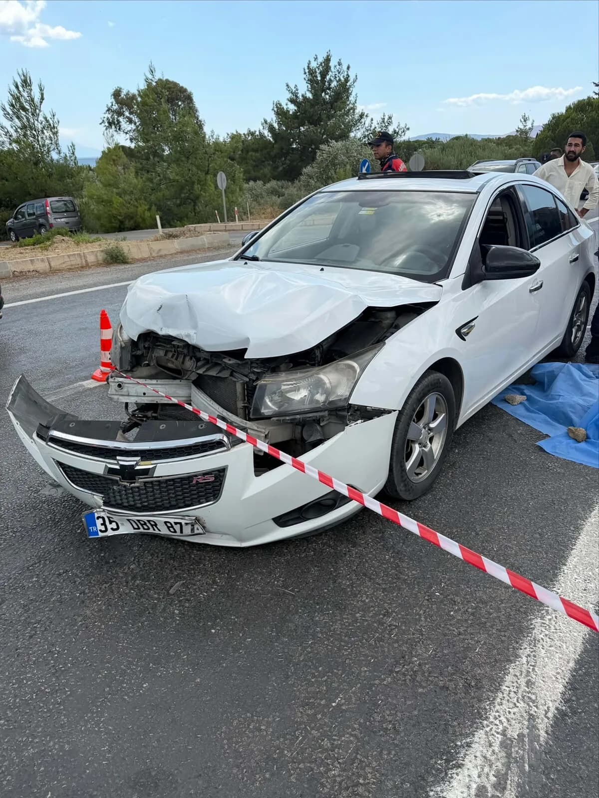Ön kısmı ve tamponu parçalanmış beyaz otomobil, yola serilmiş mavi örtü ve güvenlik şeridi