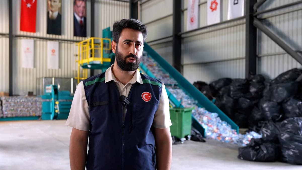 Depozito iade tesisinde üzerinde Türk bayrağı bulunan yelekli bir görevli konuşuyor, arkada preslenmiş şişeler ve dolum bandı