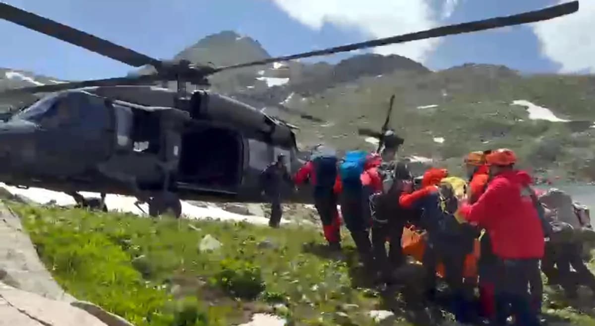 Dağlık ve karlı arazide bekleyen askeri helikopterin yanında yaralıyı sedyeyle taşıyan kırmızı montlu arama kurtarma ekipleri